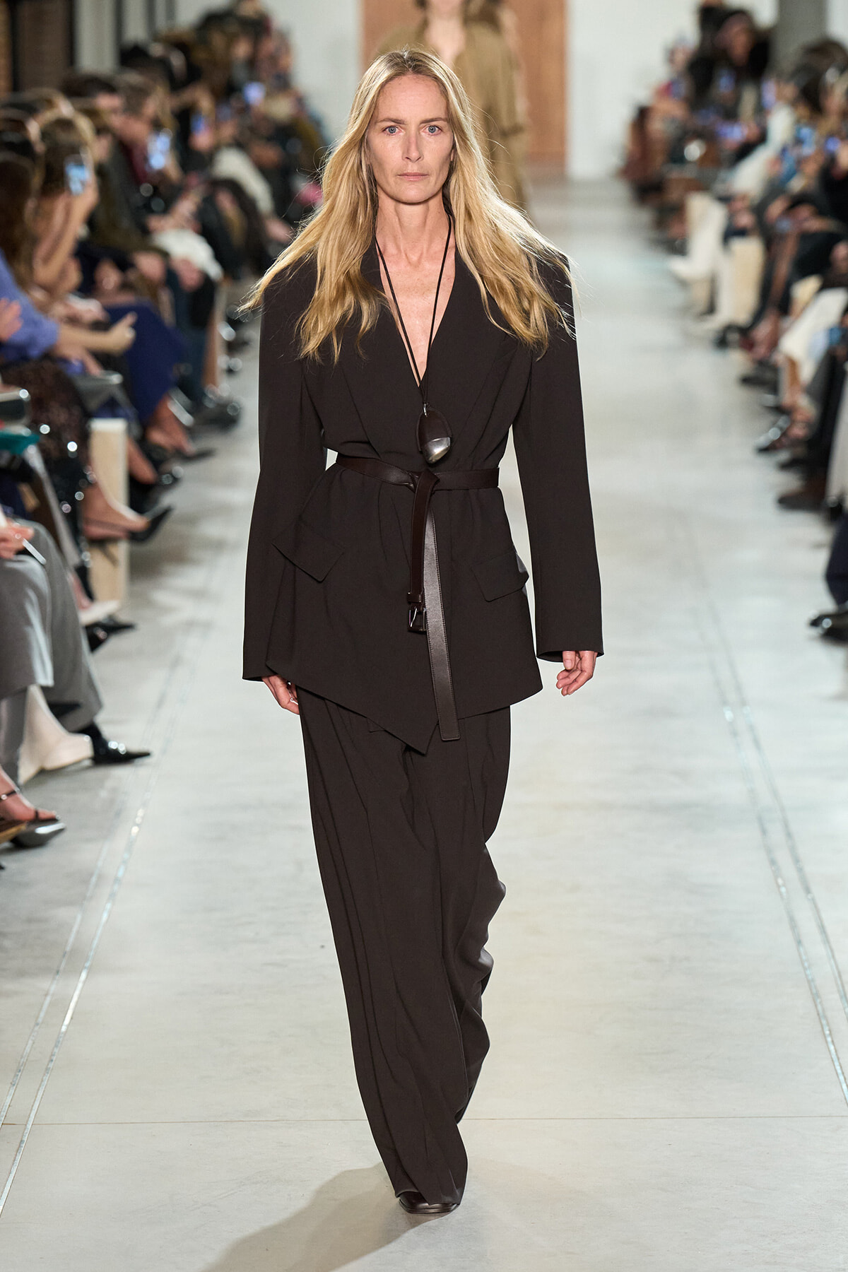 Model walks the runway wearing a black belted blazer and matching wide-leg trousers on the catwalk.