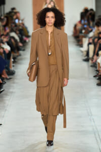 Model walking a runway in a tan coordinated suit: oversized blazer, pleated midi skirt with matching trousers, brown shirt, and a tan clutch under the arm. A long pendant necklace hangs over the shirt.