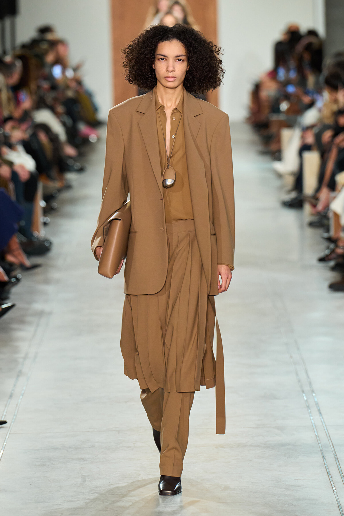 Model walking a runway in a tan coordinated suit: oversized blazer, pleated midi skirt with matching trousers, brown shirt, and a tan clutch under the arm. A long pendant necklace hangs over the shirt.