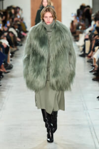 Model on runway wearing a voluminous mint-green fur coat over a gray turtleneck and beige midi skirt, with black knee-high boots.