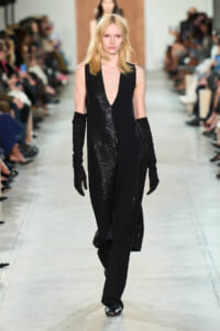 Model walks the runway in a black sequined gown with a deep V neckline and long black gloves, audience blurred in the background.