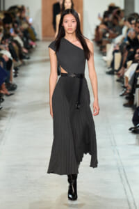 Model walking the runway in a dark gray one‑shoulder pleated dress with a black belt and tall black boots.