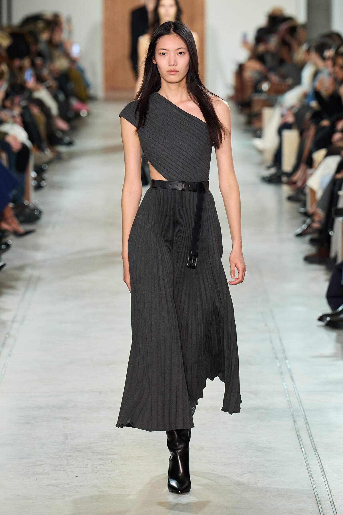 Model walking the runway in a dark gray one‑shoulder pleated dress with a black belt and tall black boots.