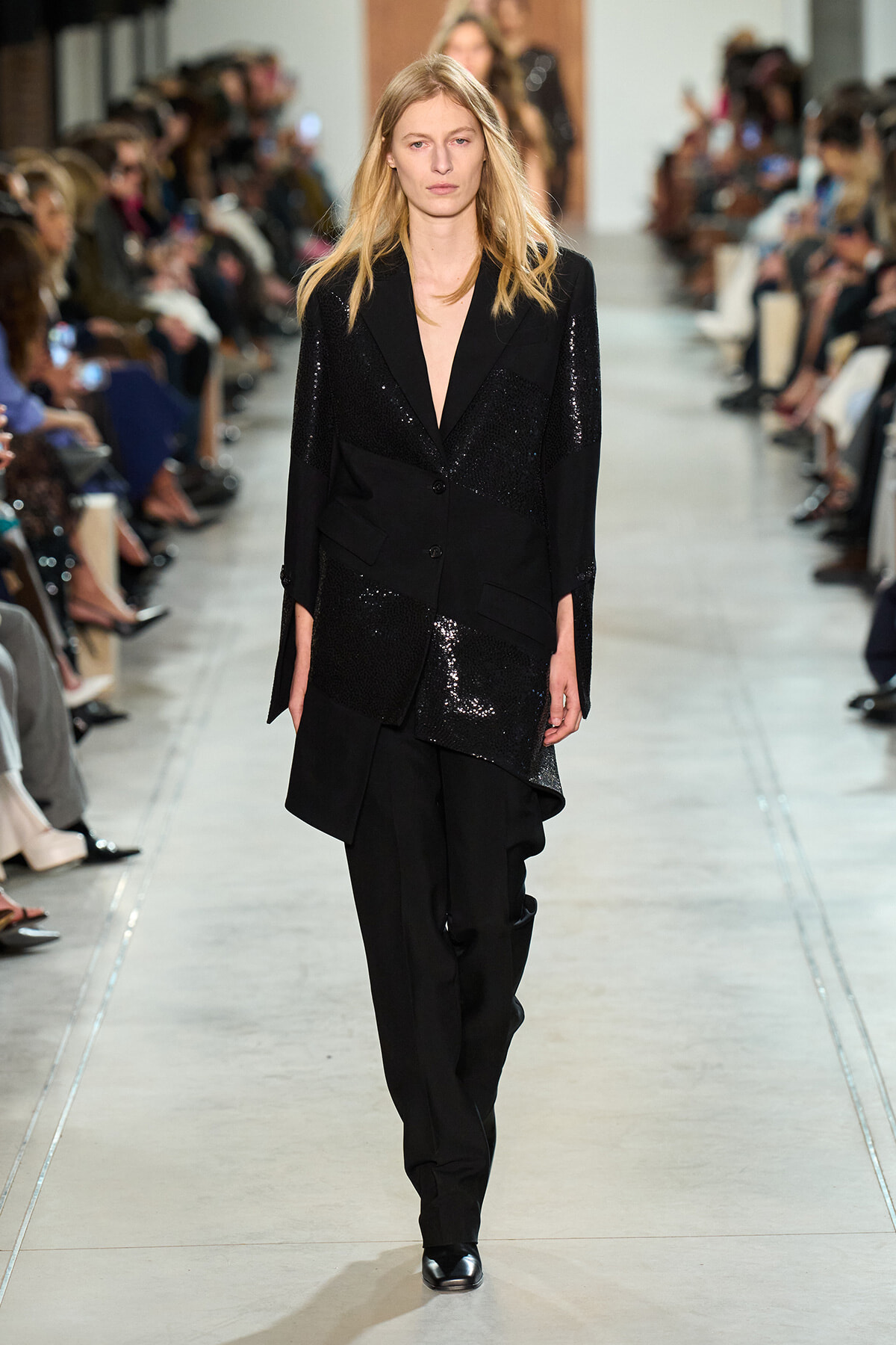 Model walks the runway in a black sequined blazer and matching pants, audience along the sides.