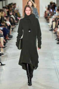 Model walks the runway in a dark green knit turtleneck dress with a matching scarf and black knee-high boots.