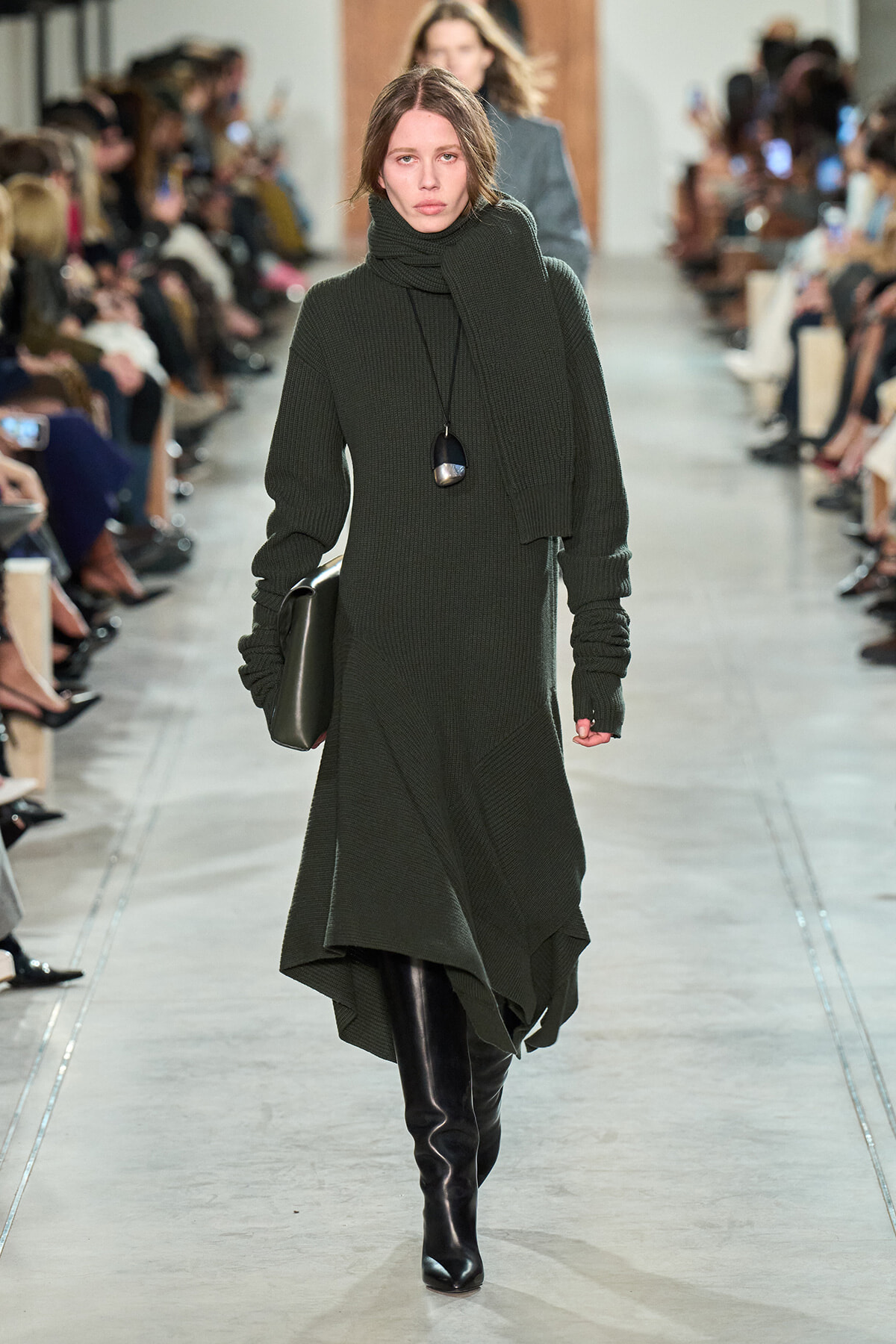Model walks the runway in a dark green knit turtleneck dress with a matching scarf and black knee-high boots.