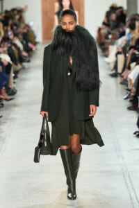 Model on a runway wearing an oversized olive-green coat with a large black fur collar, knee-high boots, and a structured gray handbag with sunglasses attached.