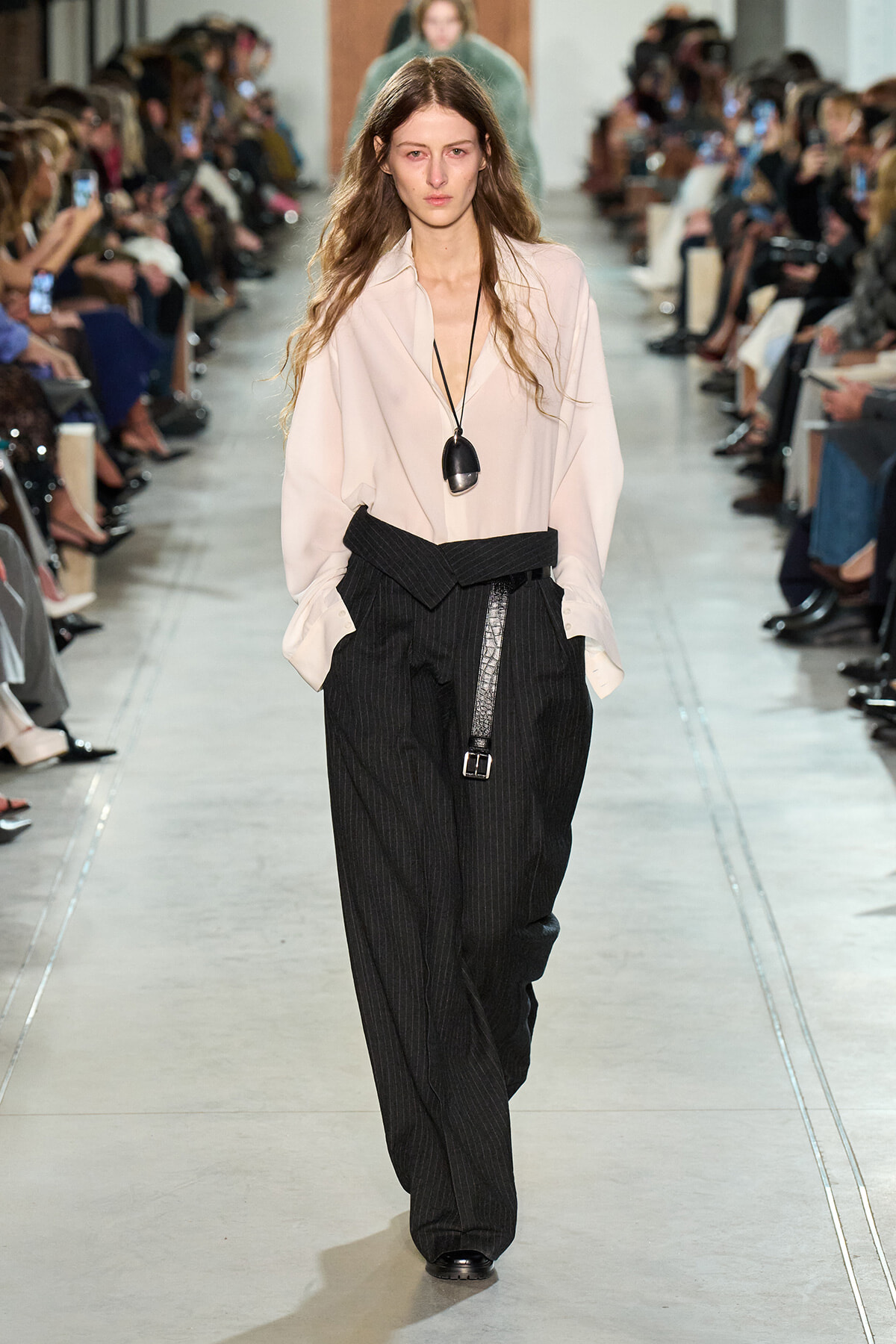 Model walks the runway in a pale pink loose blouse and oversized pinstripe black trousers with a wide belt, during a fashion show.