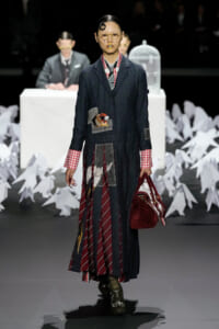 Model walks runway in a dark denim coat with patchwork pockets and a bird applique, red plaid cuffs, and a maroon striped panel skirt; carries burgundy handbag and wears round glasses.