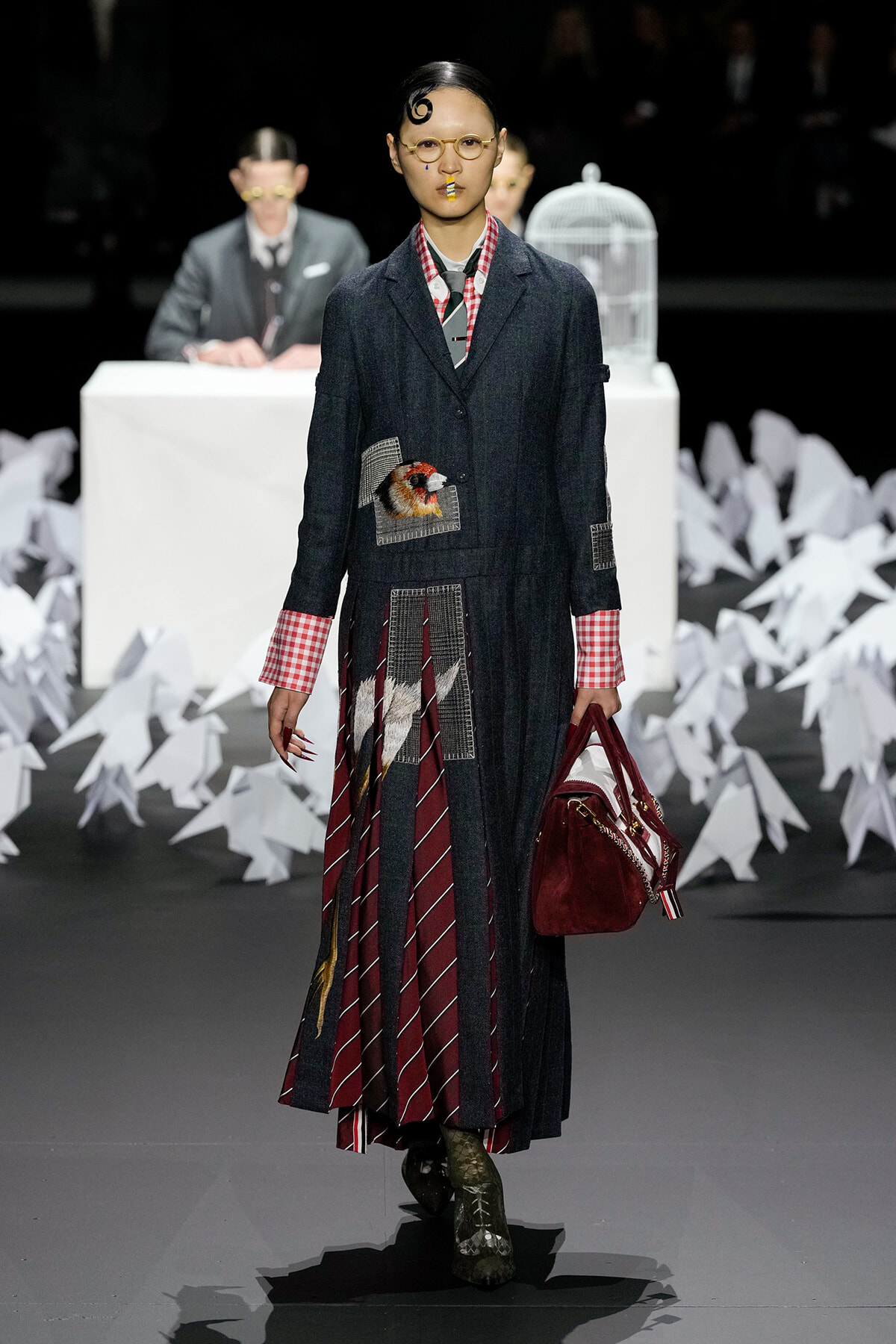 Model walks runway in a dark denim coat with patchwork pockets and a bird applique, red plaid cuffs, and a maroon striped panel skirt; carries burgundy handbag and wears round glasses.
