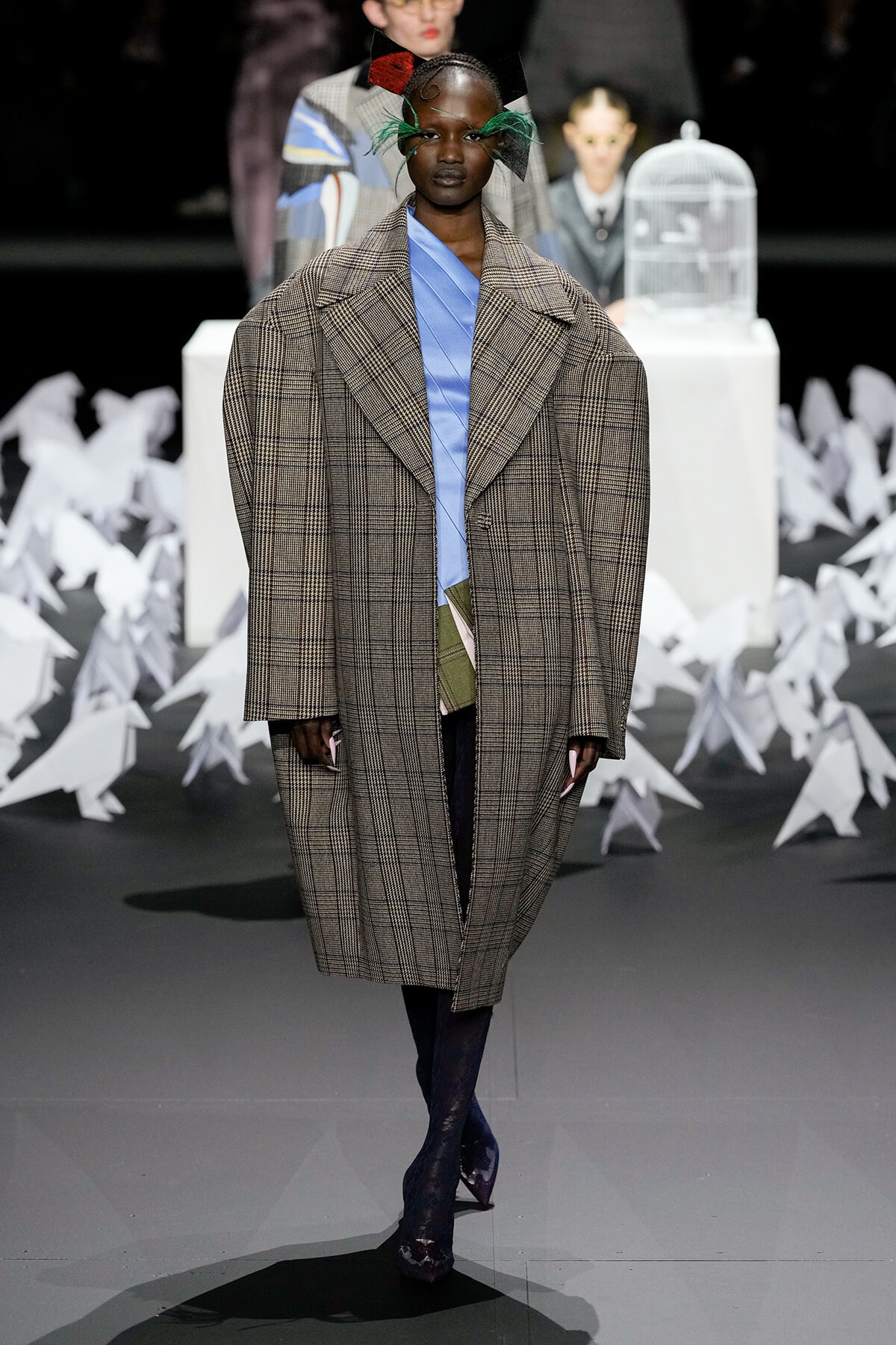 Runway model in an oversized brown plaid coat over a blue top, dark tights, and purple shoes, with a green feather headpiece; white origami birds in the backdrop.