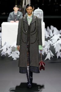 Model walks a fashion runway in a long gray tweed coat with circular black beaded trim, green gingham shirt, plaid hem, lace tights, and knee-high boots, carrying a red plush bag.