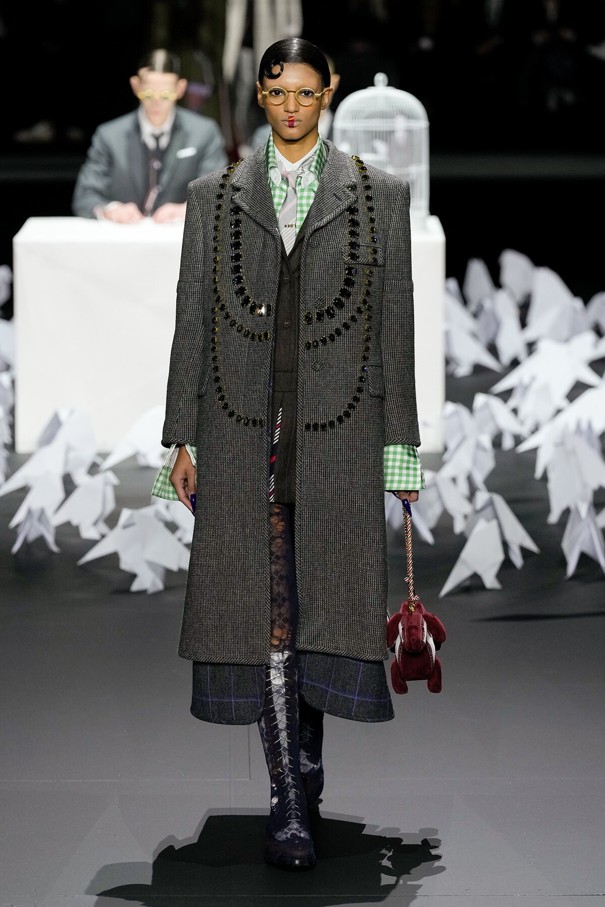 Model walks a fashion runway in a long gray tweed coat with circular black beaded trim, green gingham shirt, plaid hem, lace tights, and knee-high boots, carrying a red plush bag.