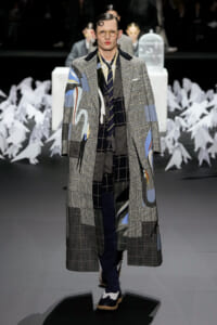 Model walks the runway in a long, patchwork plaid coat with abstract blue and yellow patches, wearing glasses and a slicked hairstyle.