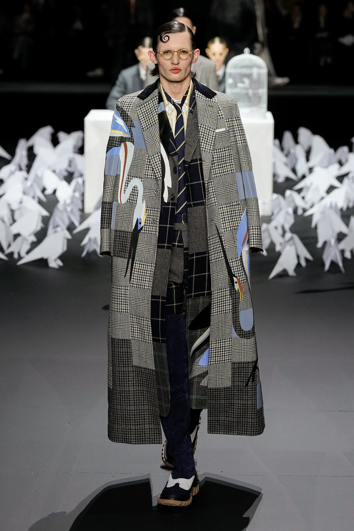 Model walks the runway in a long, patchwork plaid coat with abstract blue and yellow patches, wearing glasses and a slicked hairstyle.