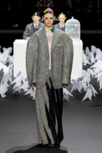 Model on runway wearing an oversized grey plaid blazer over a pink top and asymmetrical checked skirt, with a colorful headpiece.