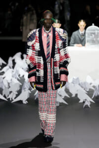 Model walks the runway in a bold patchwork knit coat with red plaid shirt and matching pants; green accessory strands in hair, dark backdrop with white sculptural shapes behind.