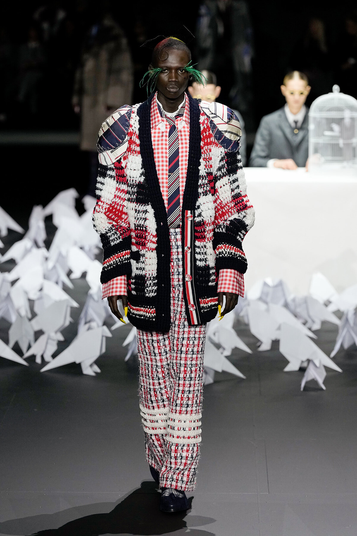 Model walks the runway in a bold patchwork knit coat with red plaid shirt and matching pants; green accessory strands in hair, dark backdrop with white sculptural shapes behind.