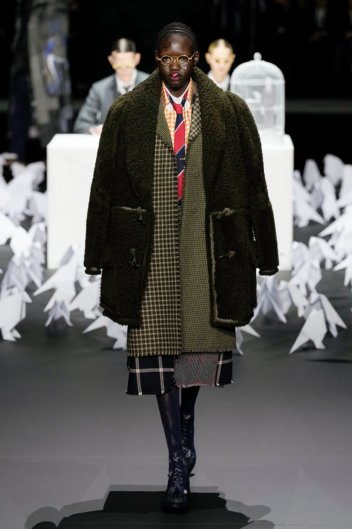 Model on a runway wearing a dark green shearling coat over layered plaid coats, round glasses, and a colorful striped tie.
