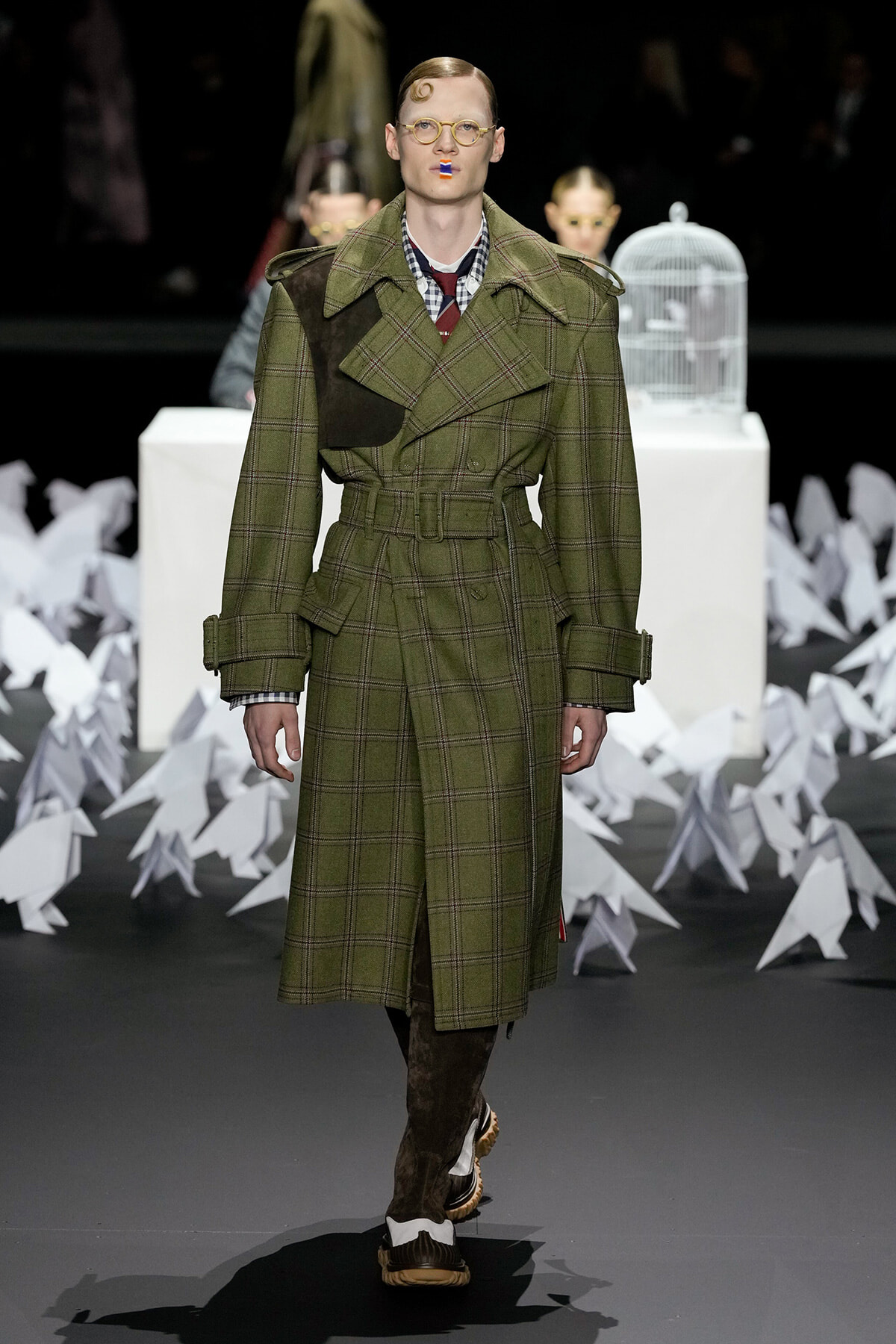 Male fashion model walking a runway in an olive plaid belted trench coat with brown suede boots and round glasses, against a backdrop of white paper shapes and a birdcage.