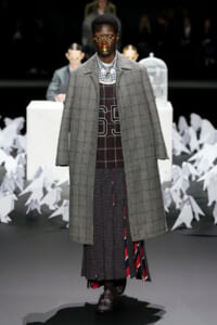 Model walking the runway in a long gray plaid overcoat over a layered knit and plaid outfit with rounded glasses and a rainbow stripe on the nose; abstract white decor in background.
