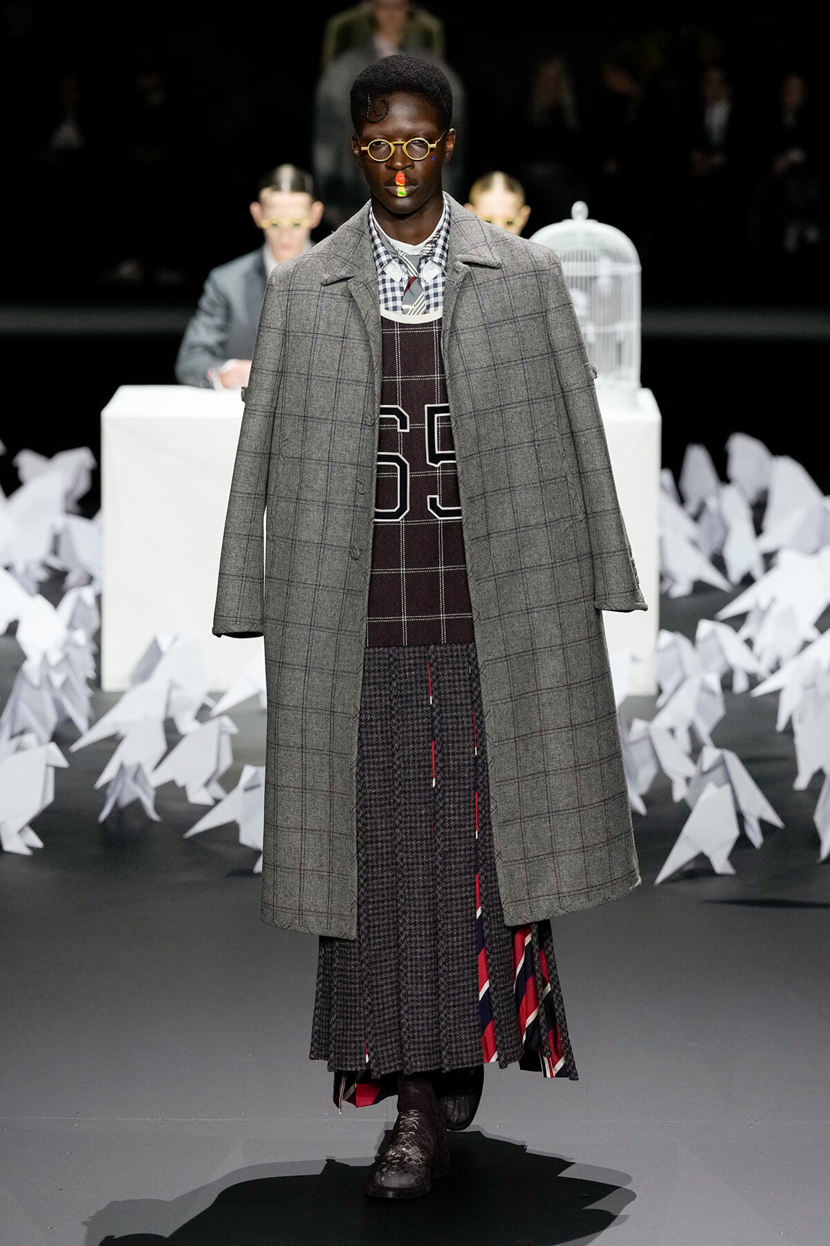 Model walking the runway in a long gray plaid overcoat over a layered knit and plaid outfit with rounded glasses and a rainbow stripe on the nose; abstract white decor in background.