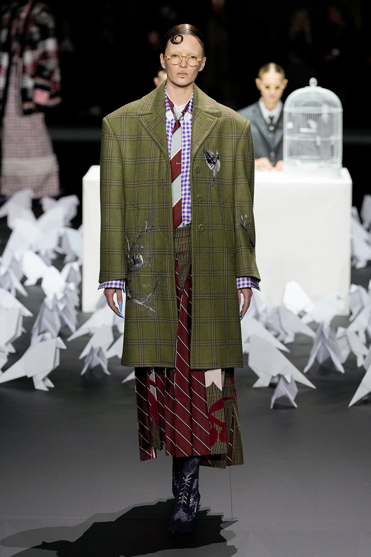 Model walks the runway in an oversized olive-green plaid coat with embroidery, layered over a patterned shirt and burgundy striped skirt; paper cranes in the background.
