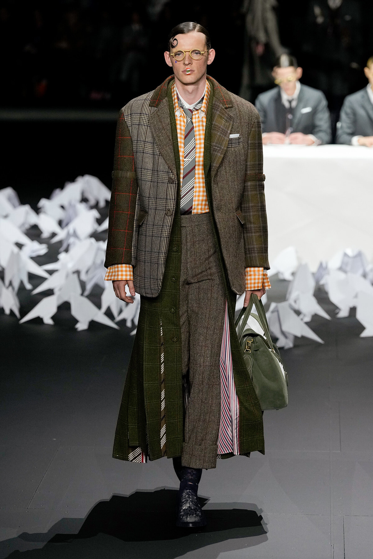 Male model walks a fashion show wearing a layered brown plaid blazer over an orange gingham shirt and striped tie, carrying a green bag.