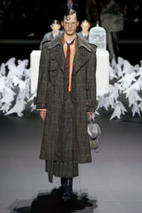 Model on a runway wearing a long plaid overcoat, orange check shirt, burgundy tie, round glasses, and carrying a gray plush backpack with a yellow key charm; paper sculptures in the background.