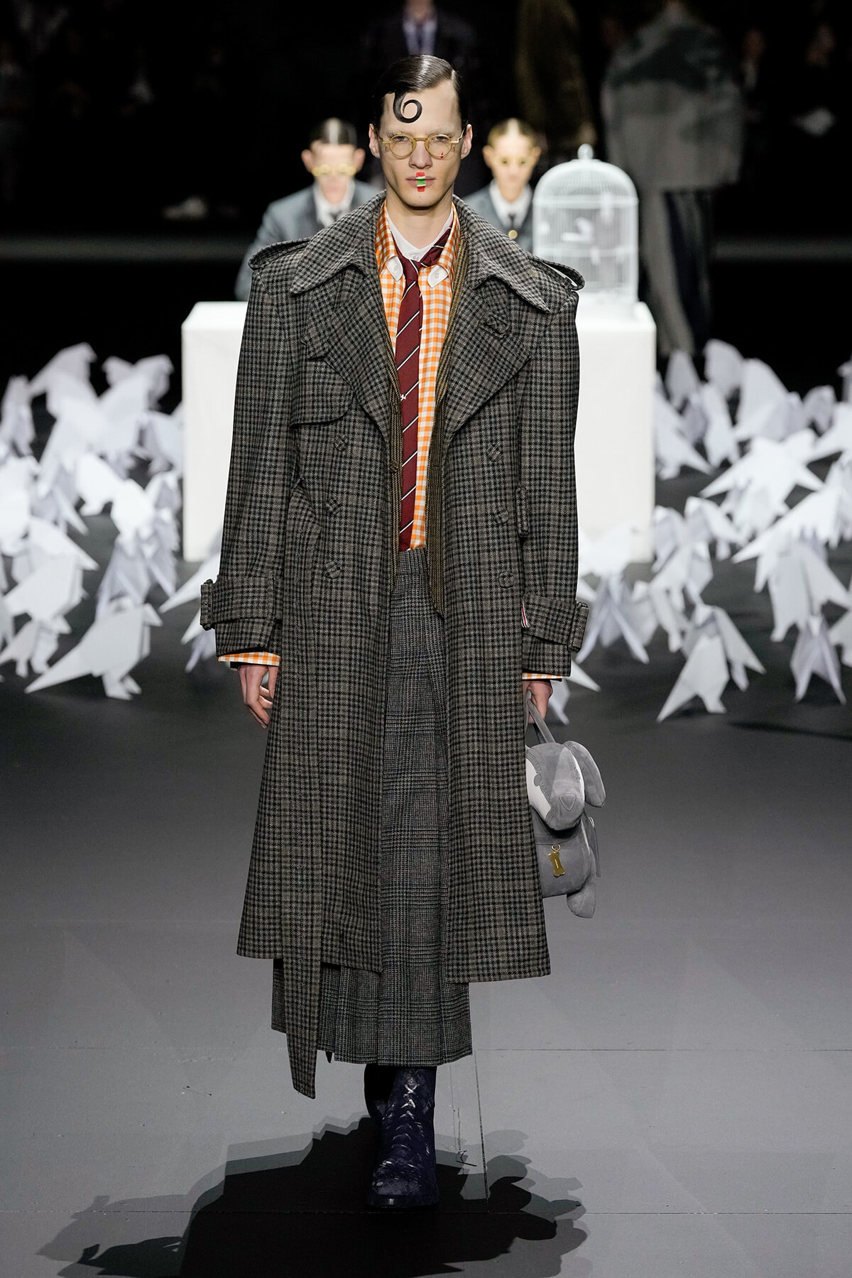 Model on a runway wearing a long plaid overcoat, orange check shirt, burgundy tie, round glasses, and carrying a gray plush backpack with a yellow key charm; paper sculptures in the background.