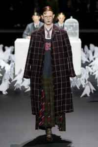 Model on a fashion show runway wearing an oversized dark plaid coat over layered plaid garments and bold yellow glasses.