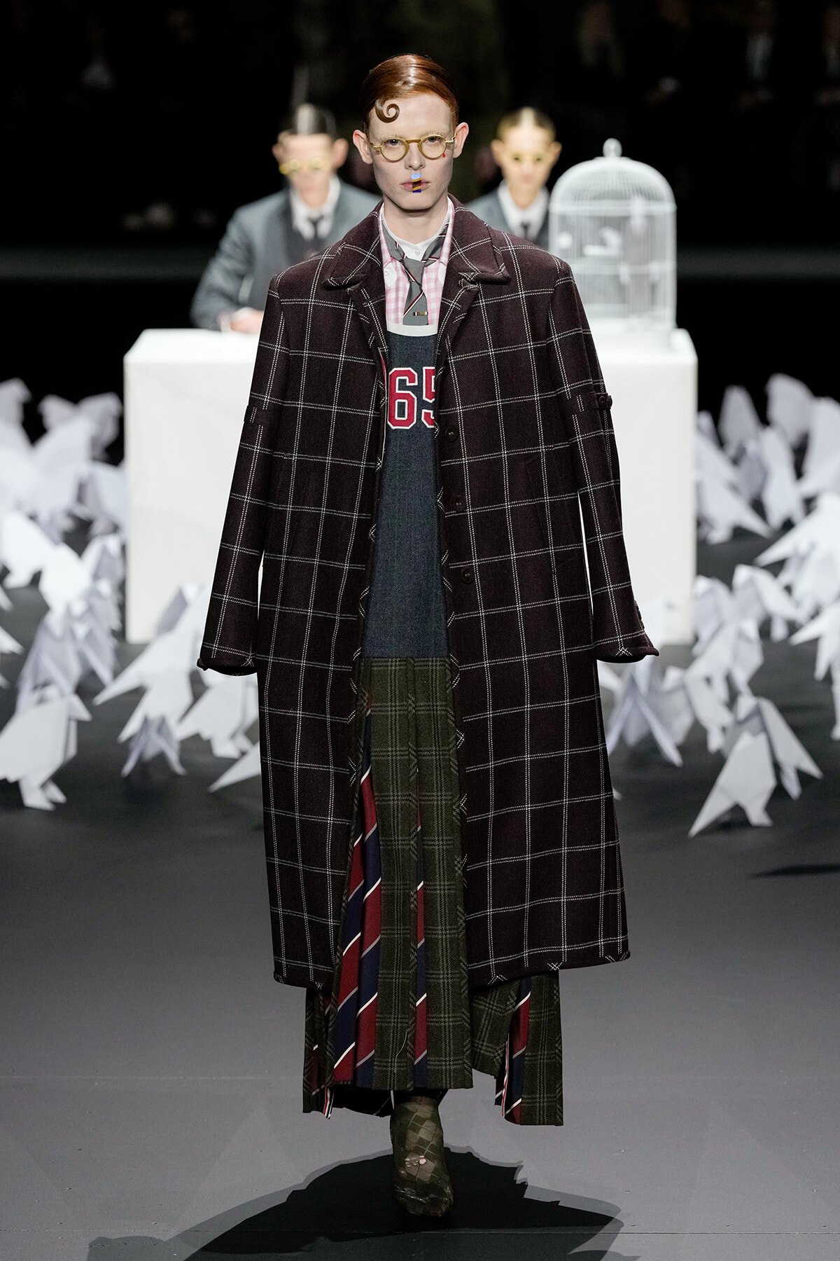 Model on a fashion show runway wearing an oversized dark plaid coat over layered plaid garments and bold yellow glasses.