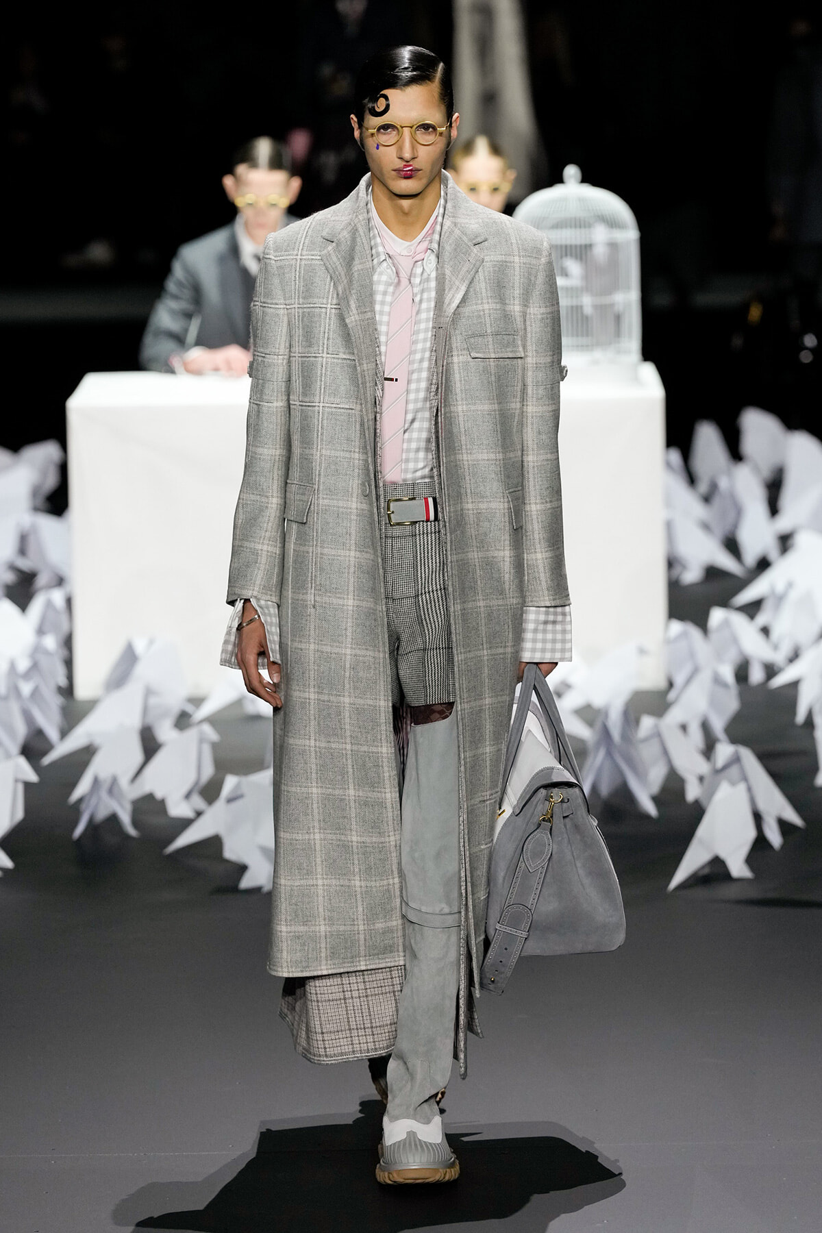 Male runway model in a long grey plaid overcoat with matching pants, pink shirt, and a grey bag on a fashion show runway with paper sculpture props in the background.