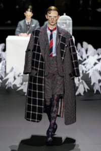 Model walks the runway in an oversized gray plaid overcoat over a matching suit, with a checkered shirt and red striped tie, plus patterned tights and round glasses.