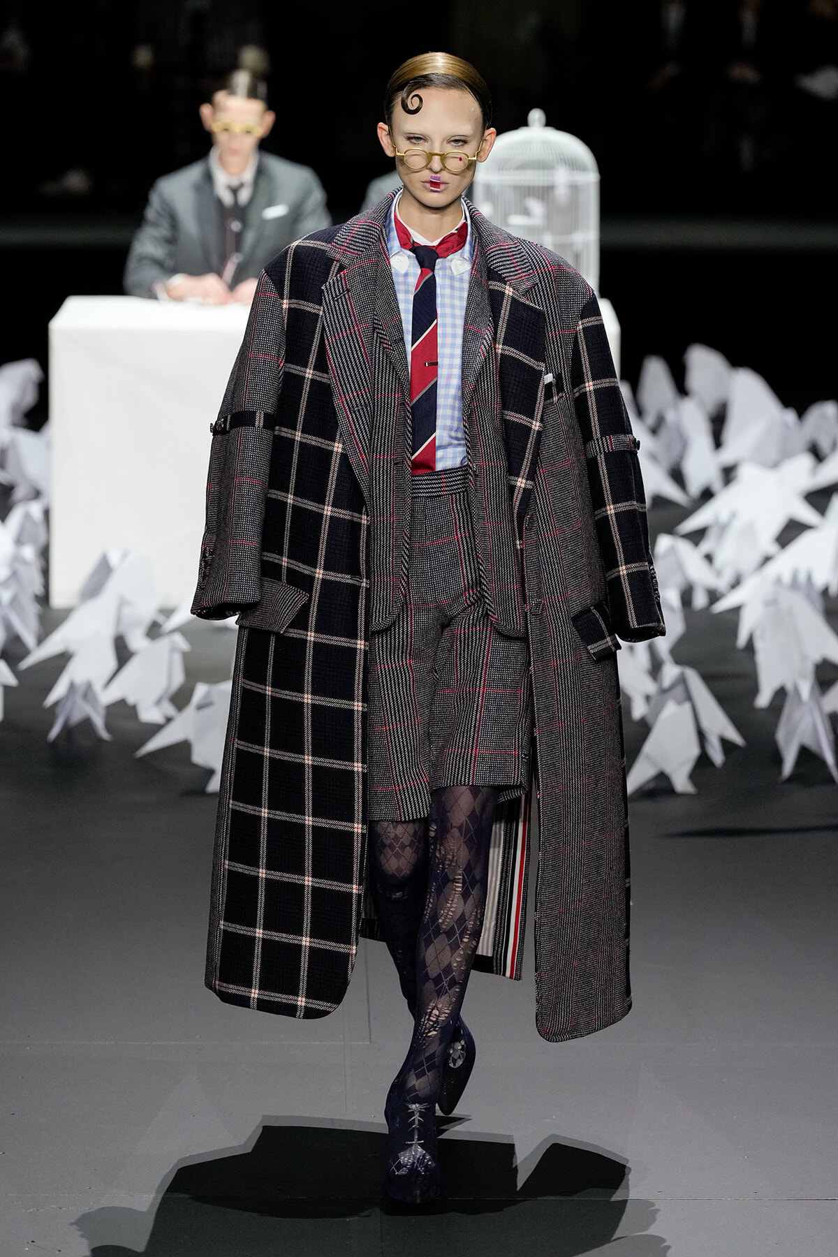 Model walks the runway in an oversized gray plaid overcoat over a matching suit, with a checkered shirt and red striped tie, plus patterned tights and round glasses.