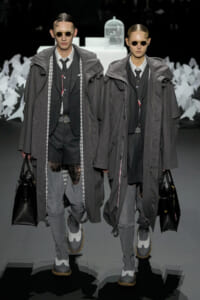 Two models walk a fashion show in matching gray layered coats, sunglasses, dark suits, and carrying black handbags.
