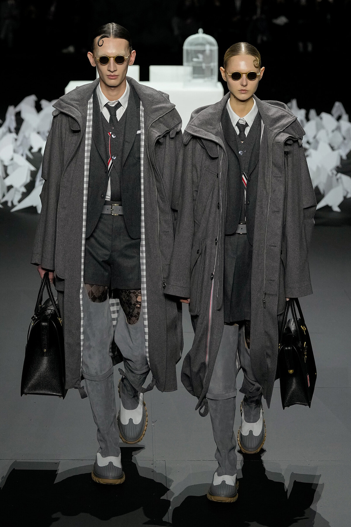 Two models walk a fashion show in matching gray layered coats, sunglasses, dark suits, and carrying black handbags.