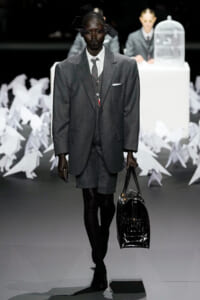 Model on a runway wearing a gray oversized suit with shorts, white shirt, and tie, carrying a shiny black handbag.