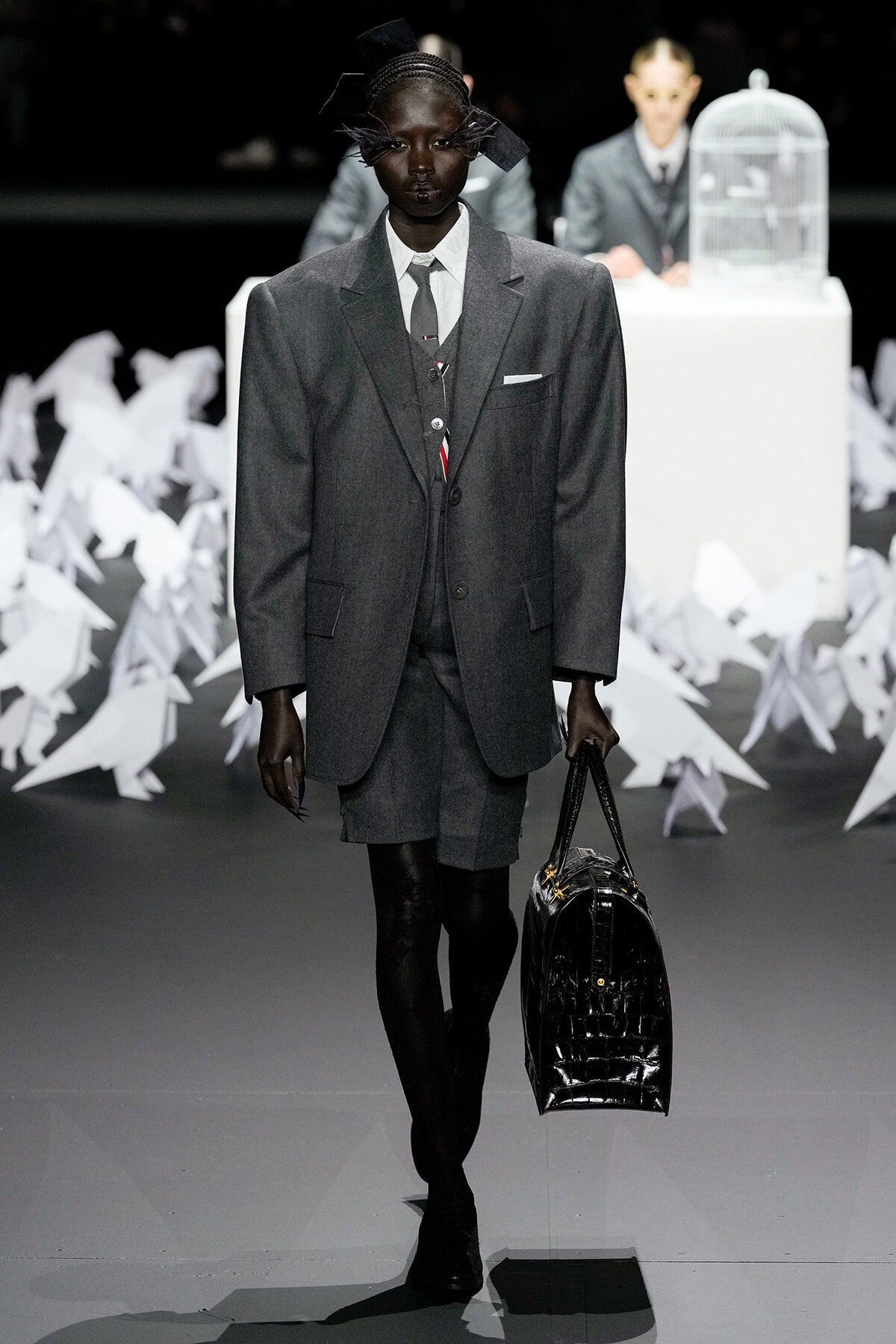 Model on a runway wearing a gray oversized suit with shorts, white shirt, and tie, carrying a shiny black handbag.