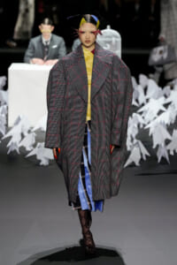 Runway model in an oversized gray plaid coat over a yellow top and blue skirt, with a bold headpiece walking forward.