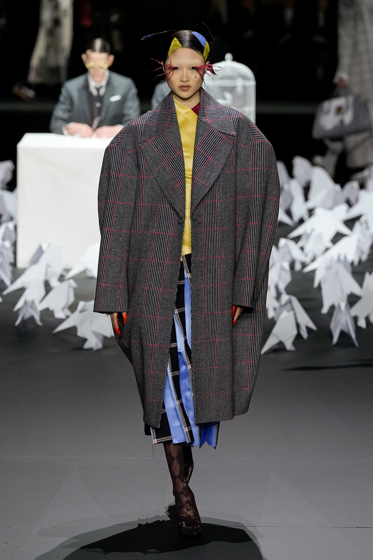 Runway model in an oversized gray plaid coat over a yellow top and blue skirt, with a bold headpiece walking forward.