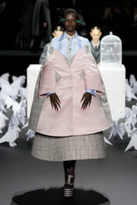 Model on a runway wearing an oversized pink satin coat with grey plaid panels over a blue gingham shirt and grey tie, bold red nails visible.