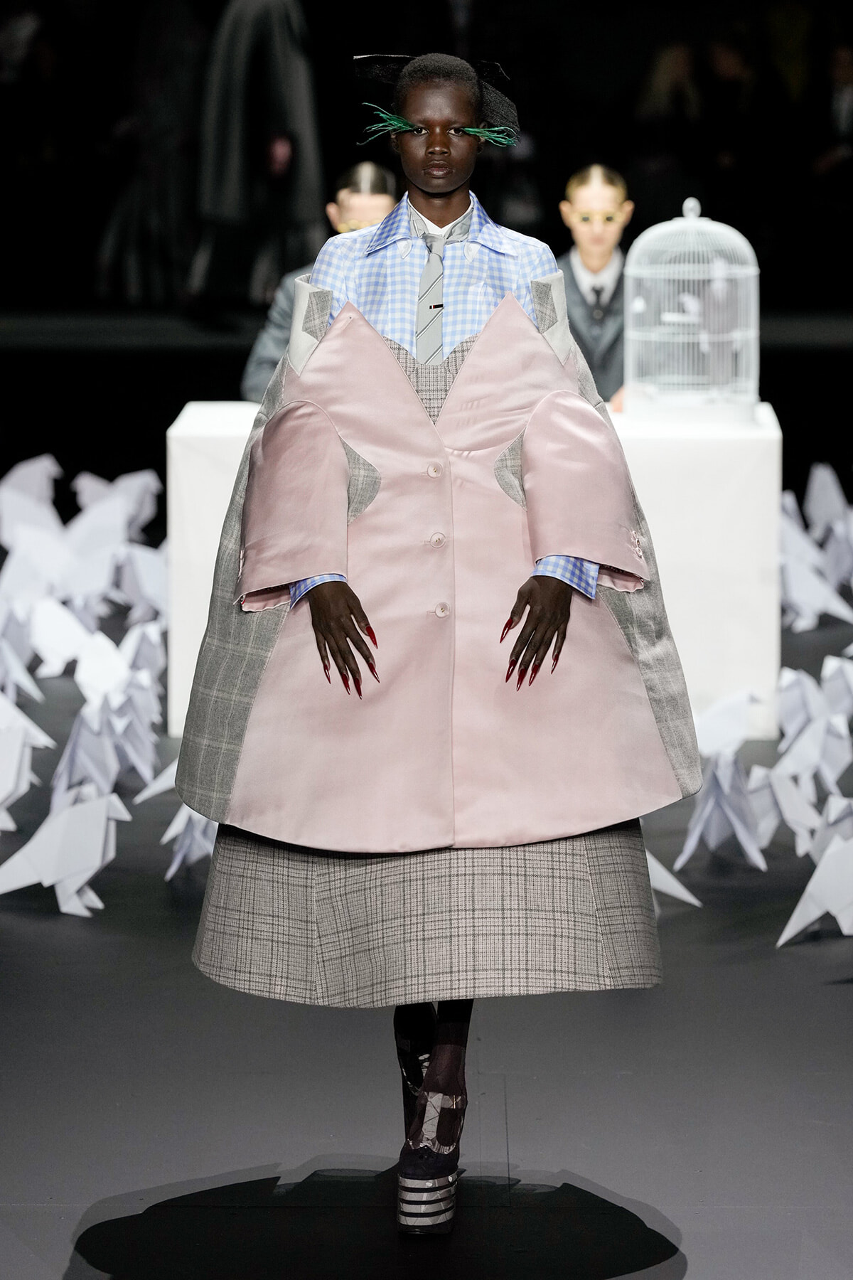 Model on a runway wearing an oversized pink satin coat with grey plaid panels over a blue gingham shirt and grey tie, bold red nails visible.