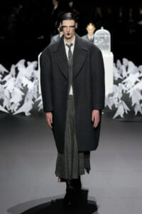 Male model on a runway wearing an oversized charcoal overcoat over a gray suit and layered skirt-like trousers, with dramatic makeup and slicked hair.