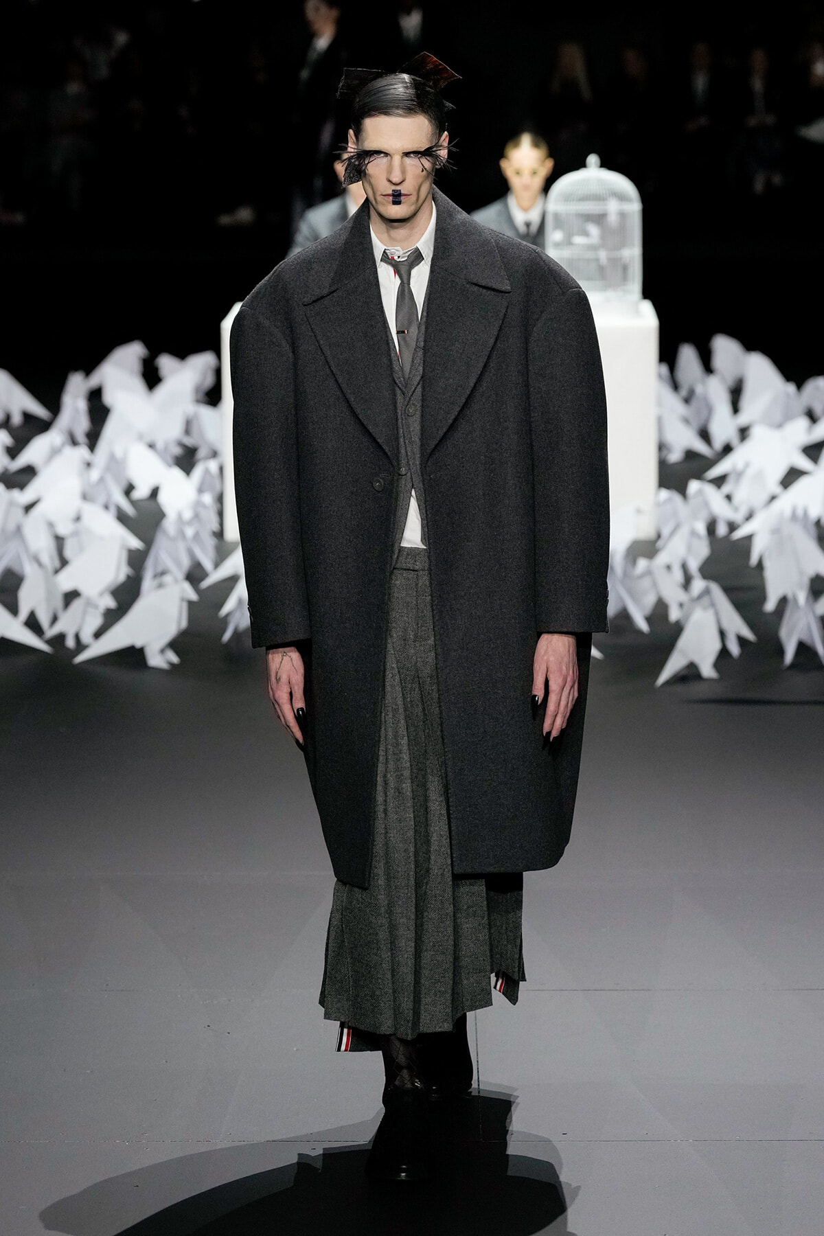 Male model on a runway wearing an oversized charcoal overcoat over a gray suit and layered skirt-like trousers, with dramatic makeup and slicked hair.