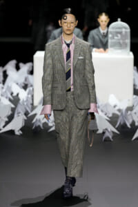 Model on a runway wearing a gray plaid suit with pink shirt cuffs, striped tie, round glasses, and a small gray handbag with a chain.