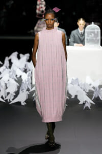 Model walks the runway in a pink plaid sleeveless midi dress with a high collar and metallic vertical seams; bold headpiece and feathered accessories.