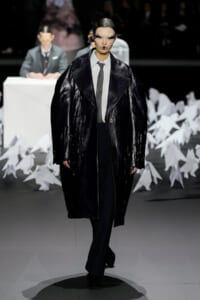 Model on a fashion runway wearing a shiny black oversized coat over a gray shirt and tie, with dramatic makeup and a bold headpiece, against a backdrop of white paper shapes on the floor.