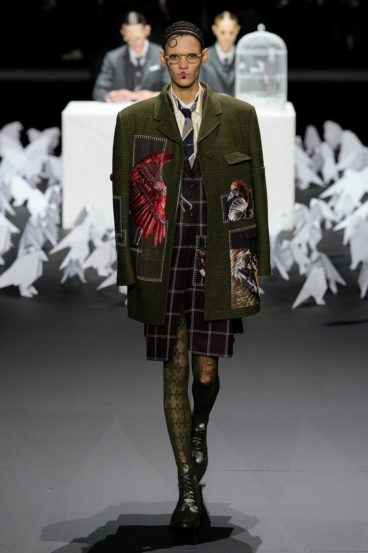 Runway model wearing a green plaid coat with bird patches over a matching plaid dress, tights, and boots, during a fashion show.