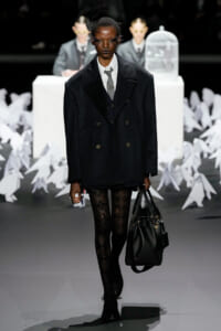 Model in an oversized double-breasted black coat, white shirt, gray tie, patterned tights, and black boots carrying a large black handbag on a fashion runway.